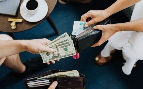 Close-up of two people exchanging US dollars and currency with wallets on a table.